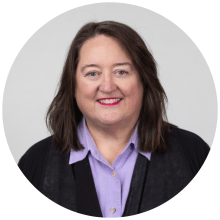 A person with brown hair smiling. They are wearing a purple shirt and a black cardigan.