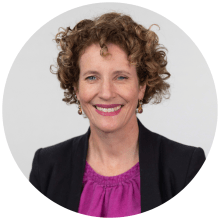 A person with curly brown hair. They are smiling and wearing a purple top and black jacket.